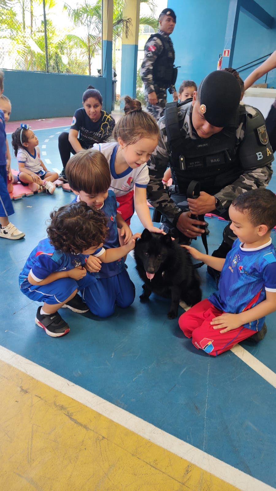 Crianças agachadas ao lado de um cão preto e de um policial, no ginásio