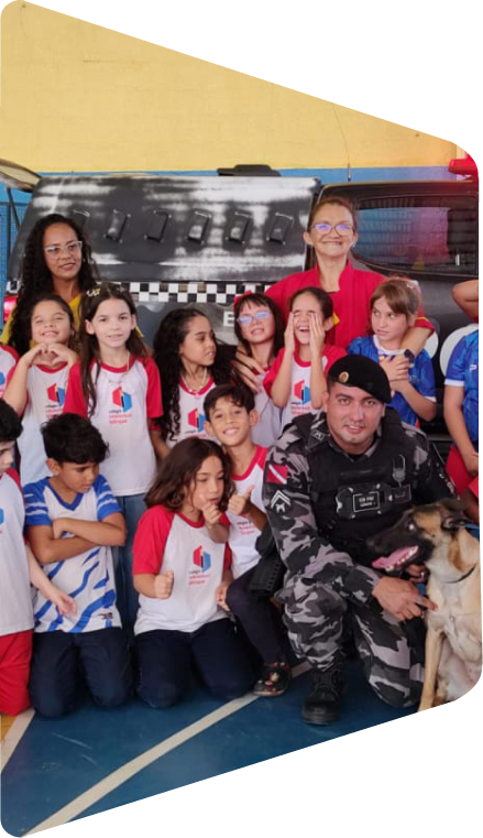 Grupo de estudantes uniformizados posando no ginásio com uma educadora, um policial e um cão